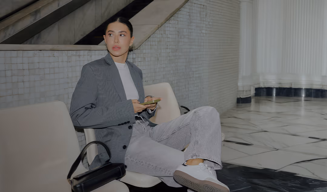Woman wearing a grey blazer and light jeans sitting on a white chair, holding a phone, with a black handbag beside her.